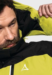 Giacca nera e verde neon con zip frontale, colletto alto e logo; presenta un tessuto testurizzato e cuciture a contrasto.