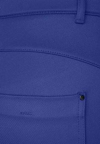 Blå denimtyg med diagonal ribbad struktur, med synliga sömmar och en liten svart metallnitar på en ficka.