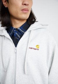 Un sweat à capuche zippé gris clair avec une texture douce. Il arbore un logo doré et bordeaux sur la poitrine ainsi que des cordons pour la capuche.