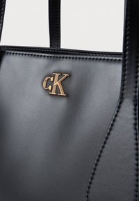 Μαύρη δερμάτινη τσάντα χειρός με λεία υφή, διαθέτει χρυσό λογότυπο 'cK' και προσεγμένα ραμμένα άκρα. Περιλαμβάνει ανθεκτικούς διπλούς ιμάντες.
