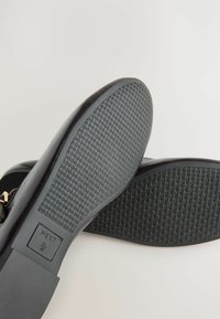 Mustad musta värvi korterjalatsite tallad, millel on tekstuuriga kummipind, millel on väike ristkülikukujuline sildike, millel on embosseeritud "NEXT" ja suuruse detailid.