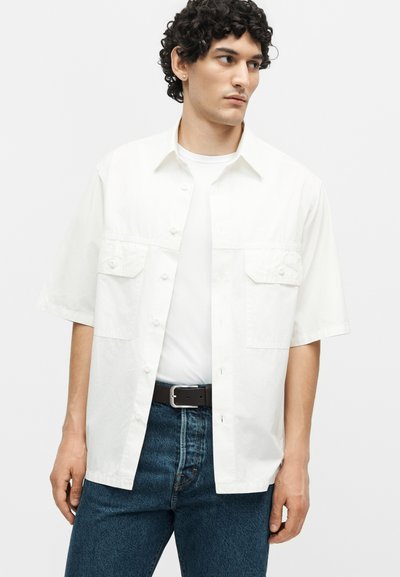 Νέος άνδρας με σγουρά μαλλιά που φοράει λευκό κοντομάνικο πουκάμισο πάνω από λευκό μπλουζάκι και μπλε τζιν με μαύρη ζώνη.