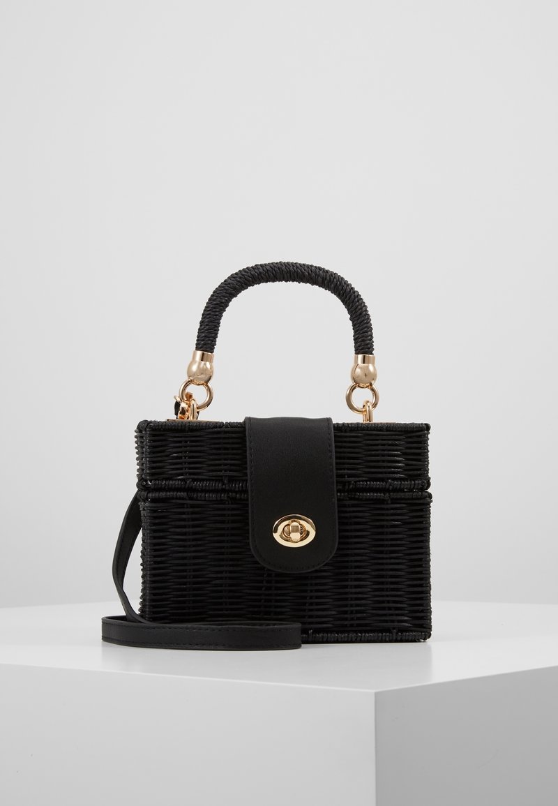 Sac à main en osier noir avec poignée tressée, fermoir doré et bandoulière amovible sur un socle blanc sur fond gris clair.