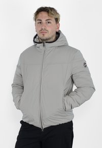 Giacca isolante grigio chiaro con cappuccio, zip frontale e tasche laterali. Presenta un piccolo logo sulla manica sinistra e polsini elasticizzati.