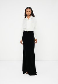 Saia maxi de veludo preto com cintura ajustada e detalhe em dourado no cinto, combinada com uma camisa branca de botão de mangas longas.