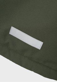 Telas de un verde oliva oscuro con una textura suave, con una etiqueta rectangular de tela blanca cosida en la parte inferior, que presenta una marca sencilla.