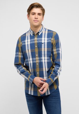 Junger Mann trägt ein blau-weiß-gelb kariertes Hemd mit langen Ärmeln und blaue Jeans, steht vor einem schlichten Hintergrund.