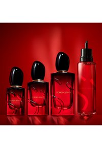 Quattro flaconi di profumo in vetro rosso con tappi neri e dettagli dorati. Il design presenta linee curve e il nome del marchio "Giorgio Armani" in oro.