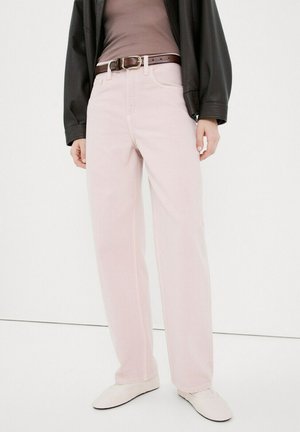 Persona vestida con pantalones rectos de color rosa claro, cinturón de cuero marrón, blusa malva, chaqueta negra y zapatos blancos sin cordones, de pie contra un fondo blanco.