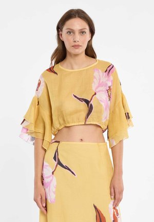 Vrouw draagt een geel tweedelig set met grote roze bloemenprints, bestaande uit een cropped top met ruches aan de mouwen en een bijpassende rok.