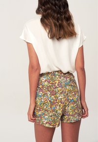 Pantaloni corti floreali con un motivo multicolore che include verdi, gialli e viola, dotati di una cintura elastica e una vestibilità comoda. Top bianco visibile.