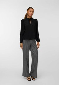 Femme aux cheveux bouclés en queue de cheval portant un blouse noire à manches longues et un pantalon large à rayures gris avec des sandales noires.