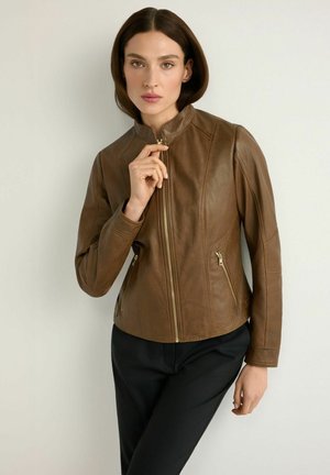 Kobieta o prostych brązowych włosach, ubrana w brązową skórzaną kurtkę z kieszeniami na zamek oraz czarne spodnie, zapinająca suwak kurtki.