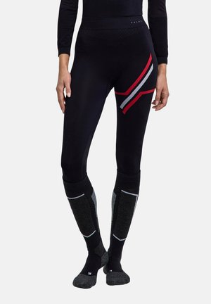 Person iført sorte leggings med røde og grå striber på låret og matchende knæhøje strømper, stående mod en ensfarvet baggrund.