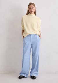 Jasnożółty sweter z dzianiny z wysokim dekoltem, w zestawieniu z szerokimi niebieskimi spodniami w paski i czarnymi butami wsuwanymi. Gładkie tło.