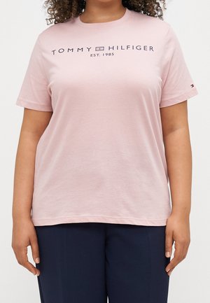 Donna che indossa una maglietta leggera rosa di Tommy Hilfiger con logo blu navy e pantaloni blu navy, in piedi con le braccia rilassate ai lati.
