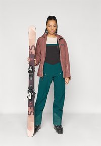 Εξοπλισμός σκι που περιλαμβάνει μια μπορντό τζάκετ, μαύρες και πράσινες φόρμες, μαύρες μπότες σκι και ένα σκι με πολύχρωμα γραφικά, κρατημένο όρθιο.