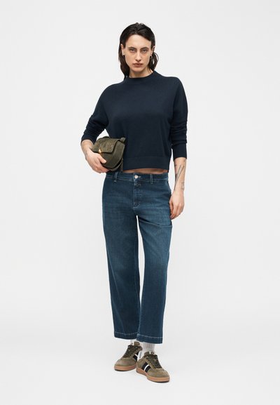 Femme debout portant un pull bleu marine, un jean taille haute bleu, des baskets vertes, tenant un sac à main vert olive sur un fond uni clair.