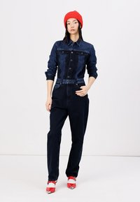 Jeans kombinezon v temno modri barvi z gumbasto zasnovo, z žepi na prsih in zategnjenim pasom. Ujemajo se z rdečimi visokimi petami in klobukom.