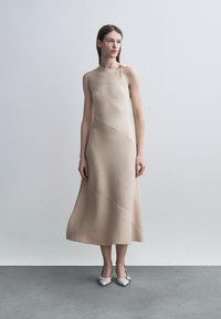 Woman stands wearing a sleeveless beige ribbed midi dress with an asymmetrical hem and pointed white heels with buckle straps.