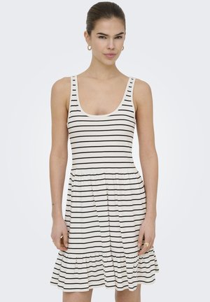 Joven mujer con vestido blanco sin mangas con finas rayas horizontales negras y dobladillo con volantes, de pie frente a un fondo claro y liso.