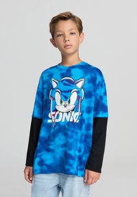 Maglietta t-shirt tie-dye blu con una grande grafica di Sonic the Hedgehog in bianco e nero, con occhi verdi, sovrapposta a una maglia a maniche lunghe nera.