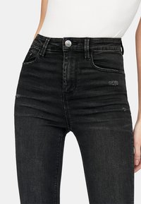 Vaqueros skinny de tiro alto en negro, hechos de denim, con un ligero desgastado, detalles desgastados, cinco bolsillos y un cierre de botón plateado.