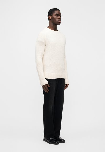 Cream-colored textured sweater with long sleeves, paired with loose-fitting dark denim jeans and black shoes, shown from the side.