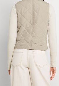 Beżowa pikowana kamizelka z wysokim kołnierzem, gładki materiał i unikalne detale szycia, noszona na jasnym swetrze ryżowym i szerokich spodniach.