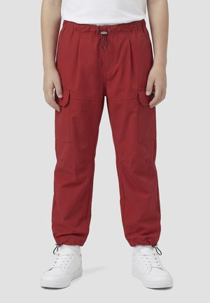 Dijete koje nosi crvene cargo hlače s elastičnim manšetama i vezicom u struku, uparene s bijelim tenisicama i bijelom košuljom.