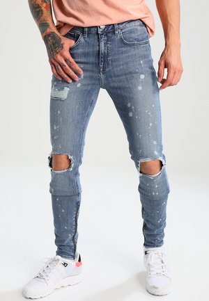 Hombre con vaqueros pitillo desgastados de color azul claro con rodillas rotas, detalles de cremallera en los tobillos, zapatillas blancas y camisa melocotón.