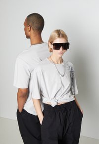 Szürke, oversized pólók, az egyik megkötve, modelleken viselve. Fekete, bő nadrágok és napszemüveg is látható. Egyszerű, letisztult vonalak minimális részletekkel.