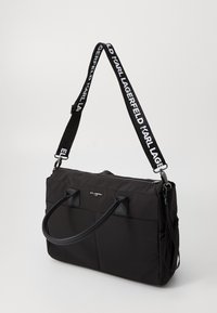 Zwarte nylon draagtas met dubbele handgrepen en een afneembare schouderriem met een logoband. Gladde textuur, ritssluiting, minimalistisch ontwerp.