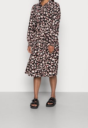 Nainen, jolla on päällään pitkähihainen, napitettava leopardi-kuosinen mekko, jossa on solmittava vyötärövyö, sekä mustat avokkaat vaaleaa taustaa vasten.