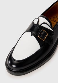 Mocasín de cuero en blanco y negro con una correa de hebilla dorada sobre un fondo blanco, mostrando los detalles frontales y laterales del zapato.