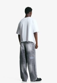 Biała, oversize koszulka z krótkim rękawem. Szare, szerokie dżinsy z kieszeniami i jasnoniebieskim logo po lewej stronie uda. Czarne obuwie.