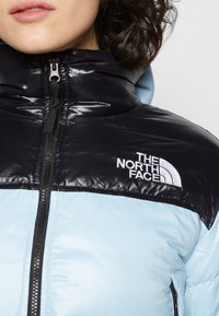Žena má na sobě světle modrou a černou péřovku s černým zipem a logem The North Face na hrudi.