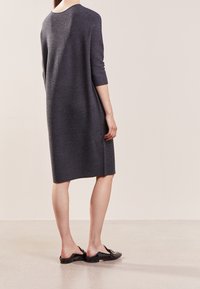 Donna che indossa un vestito a maglia grigio scuro fino al ginocchio con maniche a tre quarti e mocassini neri senza lacci, in piedi su un pavimento chiaro.