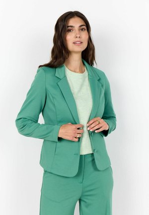 Hosszú sötét hajú nő zöld blézert és hozzáillő nadrágot visel, és a blézer gombjait igazítja egy egyszerű, fehér háttér előtt.