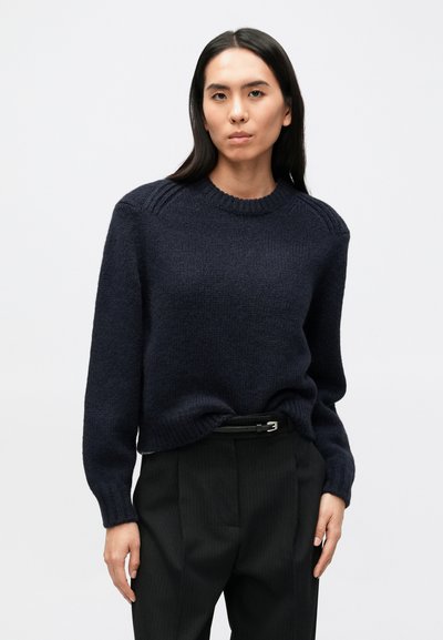 Pull en maille bleu marine avec col rond, poignets côtelés et détails aux épaules, porté avec un pantalon noir plissé et une ceinture. Texture lisse.