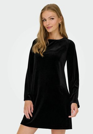 Jeune femme aux longs cheveux blonds portant une robe noire en velours à manches longues, debout devant un fond clair uni.