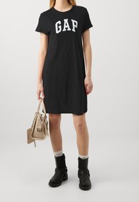 Černé tričkové šaty s bílým logem "GAP", krátké rukávy, volný střih. V kombinaci s béžovou kabelkou a černými kotníkovými botami se sponami.