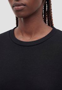 Primer plano de una persona con piel oscura y cabello trenzado que lleva una camiseta negra de cuello redondo sobre un fondo claro y liso.