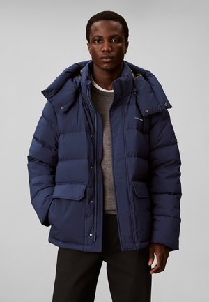 Giovane uomo che indossa una giacca imbottita blu navy con collo alto e tasche con patta, maglione grigio e pantaloni neri, in piedi davanti a uno sfondo semplice.