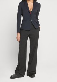 Blazer marine à coupe cintrée, col à revers nets, fermeture à un seul bouton et deux poches avant ; associé à un pantalon noir à jambes larges et des chaussures plates.