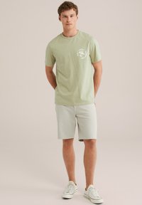 Helles grünes Baumwoll-T-Shirt mit Rundhalsausschnitt und einem weißen Grafikdruck auf der Brust. Kombiniert mit hellgrauen Shorts und weißen Sneakers.