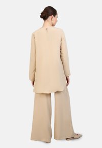 Beige tuniek met lange mouwen, hoog-laag zoom en een sleutelgatdetail aan de achterkant, gecombineerd met brede beige broek en puntige flats.