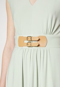 Svěže zelené šaty s plisovanou suknią a širokým opaskem ze světlého umělého kůže, který má dvě zlaté spony. Měkká látka s hladkou strukturou.