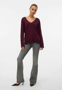 Maglione bordeaux a V con maniche lunghe, indossato con jeans a zampa d'elefante grigio chiaro e scarpe con tacco marrone, caratterizzato da un design semplice e una texture liscia.