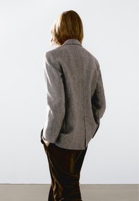 Persona con capelli di lunghezza spalle, che indossa un blazer grigio e pantaloni scuri, in piedi con le mani nelle tasche, di fronte a uno sfondo bianco semplice.